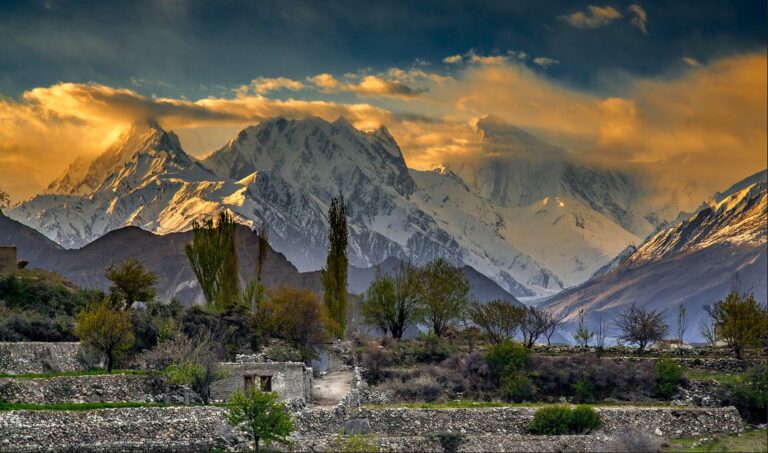 A breathtaking landscape showcasing a mountain range with peaks partially covered in snow, illuminated by the warm glow of the setting sun. The sky is painted with hues of orange and yellow, contrasting against the cool tones of the mountains. In the foreground, there are terraced fields and tall trees, with a small stone structure. The scene captures the serene beauty of nature’s grandeur at dusk.